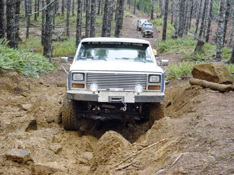 1985 Ford Bronco - A group of Ford Broncos tackle a challenging off-road course in a wooded area.