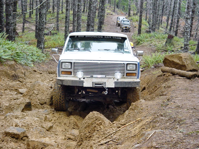 White Ford Bronco navigating a muddy trail with other vehicles behind it