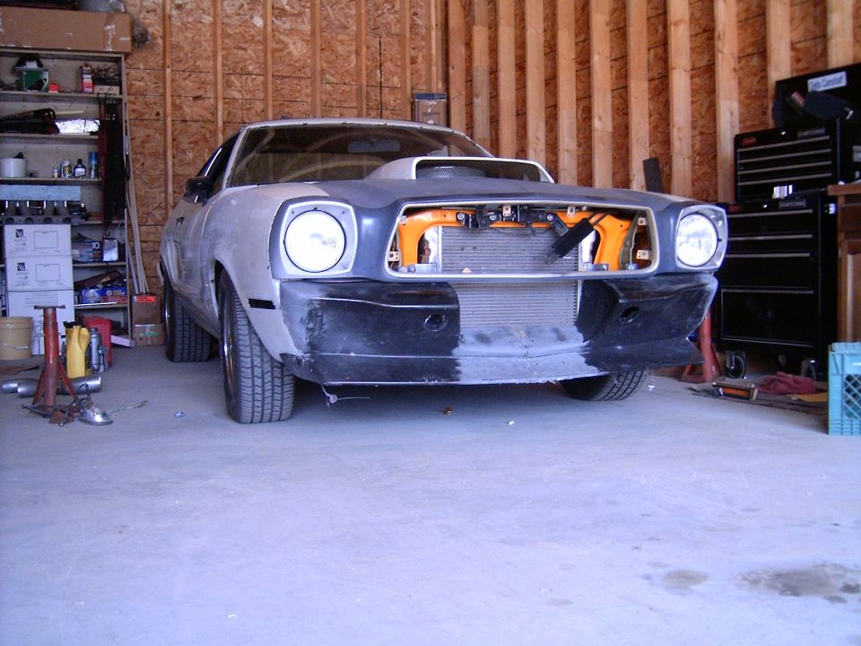 Front view of a 1978 Ford Mustang II in a garage, undergoing restoration with the hood open and bumper removed