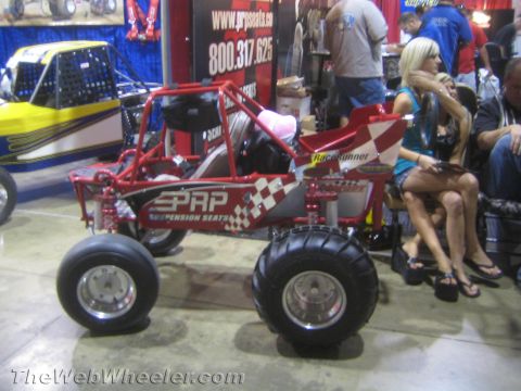 1990 Ford Bronco - red tube-frame off-road buggy with PRP suspension-seat graphics and paddle tires displayed on carpet at an indoor expo.