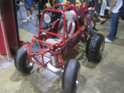 1990 Ford Bronco - small red tube-frame buggy with slick front and paddle rear tires parked on a polished show floor with onlookers behind.