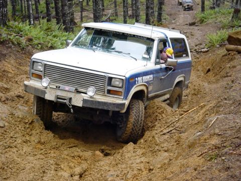 1985 Ford Bronco - A Ford Bronco navigates a challenging muddy off-road course, with another vehicle visible in the background.