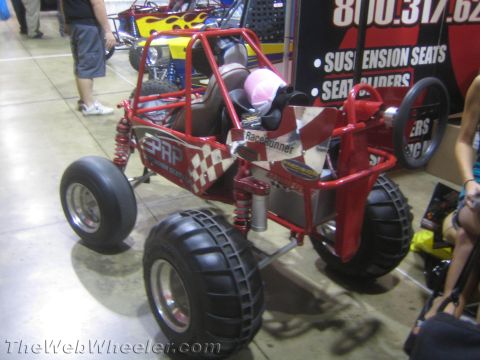 1990 Ford Bronco - red mini tube-frame buggy with checkered graphics and 'RaceRunner' lettering parked beside a PRP suspension-seats booth.