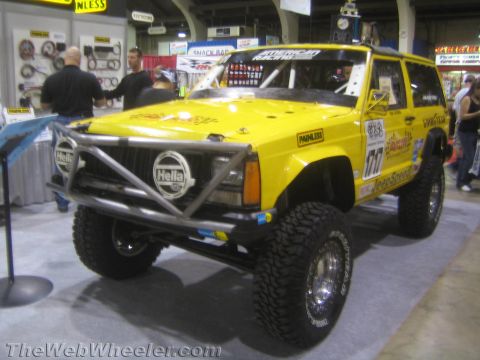 1990 Ford Bronco - yellow lifted Jeep Cherokee with a tube front bumper, Hella lights, sponsor decals and large mud tires on display indoors.