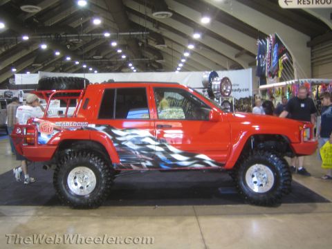 1990 Ford Bronco - Side profile of a red lifted Jeep Grand Cherokee with tribal flame graphics, roof spare tire and beadlock wheels at an indoor show.
