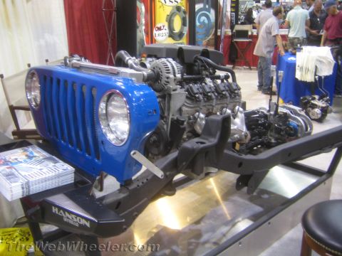 1990 Ford Bronco - blue Jeep front grille and clip mounted on a bare frame with an exposed V8 engine and Hanson tube bumper displayed at an expo.