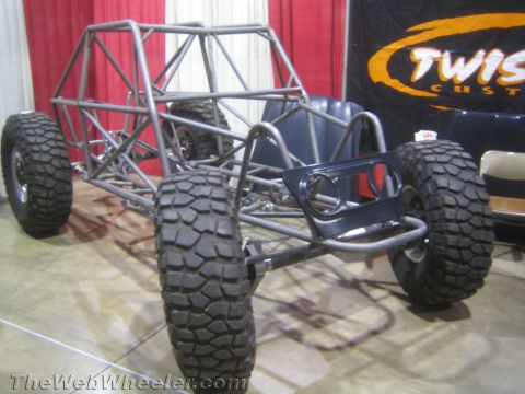 1990 Ford Bronco - bare gray tube-frame buggy chassis with large mud tires displayed in front of a Twisted Customs banner at an indoor show.