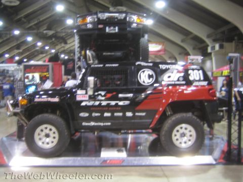 1990 Ford Bronco - black and red Toyota off-road race truck numbered 301 with KC lights and Nitto graphics displayed on a lit platform.