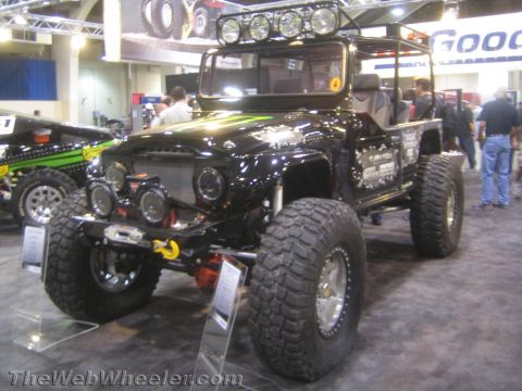 1990 Ford Bronco - black FJ40-style Toyota Land Cruiser pickup, lifted on large mud tires with a roof light bar, displayed at an indoor expo.
