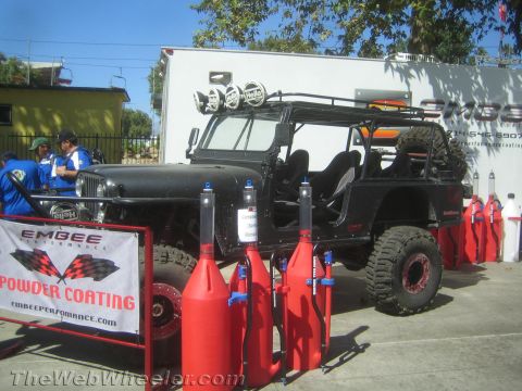 1990 Ford Bronco - black FJ40-style truck with a roof rack and light bar parked outdoors behind an Embee Powder Coating banner and red gas jugs.