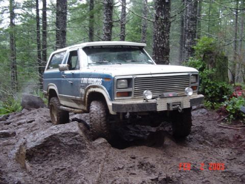 1985 Ford Bronco - This Ford Bronco is navigating a challenging off-road trail with muddy terrain and large rocks.