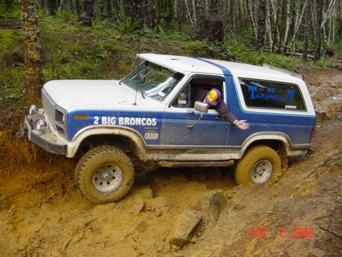 1985 Ford Bronco - A Ford Bronco navigates a muddy off-road trail, with a person's arm visible from the driver's side window.