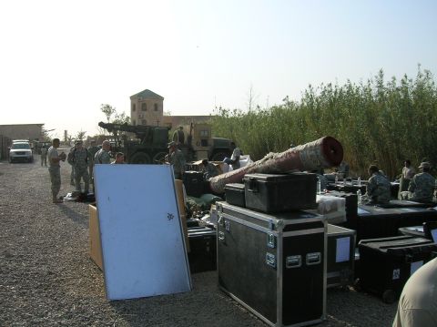 1996 Ford Bronco - Soldiers stand near a large artillery piece and various storage cases, with buildings and vegetation in the background.