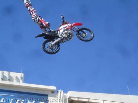 1990 Ford Bronco - freestyle motocross rider hangs off a red dirt bike mid-jump against a clear blue sky above a building.