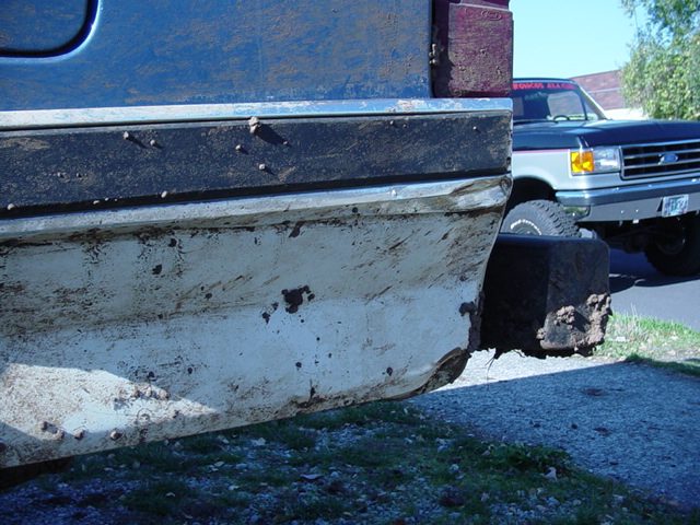 Rusty white rocker and lower door of blue truck with mud flap, Ford pickup parked behind