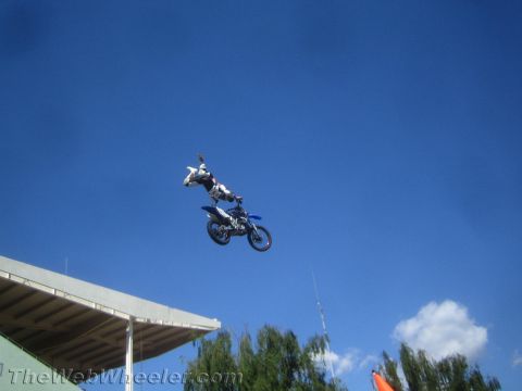 1990 Ford Bronco - freestyle rider clicks his heels over a blue dirt bike mid-jump above a stadium roof and trees.