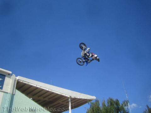1990 Ford Bronco - freestyle motocross rider performs a trick on a blue dirt bike high above a stadium roof and trees.