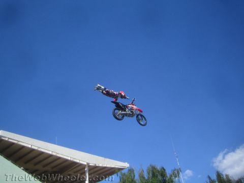 1990 Ford Bronco - freestyle rider extends off a red dirt bike during a high jump above a stadium roof and trees.