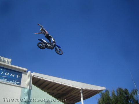 1990 Ford Bronco - freestyle motocross rider stretches off a blue dirt bike during a high jump above a stadium roof.