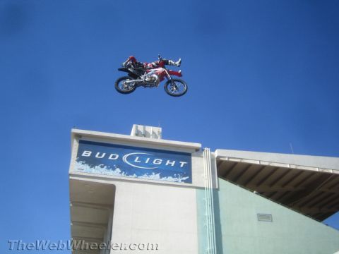 1990 Ford Bronco - freestyle motocross rider launches a red and white dirt bike high above a stadium roof bearing a Bud Light sign against a blue sky.
