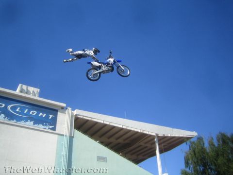 1990 Ford Bronco - motocross rider stretches fully extended off the back of a blue dirt bike in a superman trick high above a stadium against a clear blue sky.