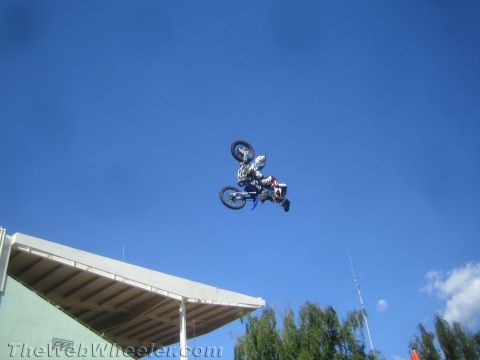 1990 Ford Bronco - motocross rider rotates through a backflip on a dirt bike high in the air above a stadium roofline and blue sky.
