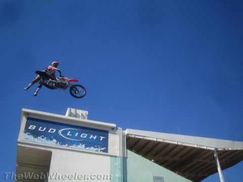 1990 Ford Bronco - motocross rider throws a red dirt bike sideways in a whip high above a stadium roof with a Bud Light sign against a blue sky.