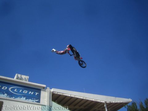 1990 Ford Bronco - motocross rider hangs inverted on a dark dirt bike at the peak of a freestyle jump high against a deep blue sky over a stadium.