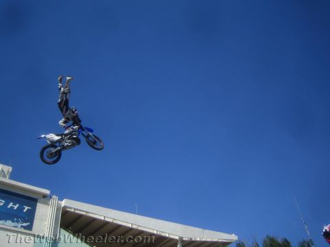 1990 Ford Bronco - motocross rider extends into a near handstand off a blue dirt bike at the top of a freestyle jump against a clear blue sky.