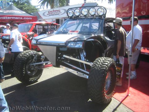 1990 Ford Bronco - black tube-chassis off-road buggy with a roof light bar and large mud tires sits on display under vendor tents at an outdoor show.