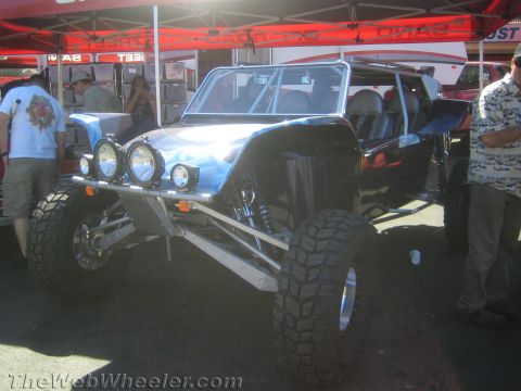 1990 Ford Bronco - blue tube-chassis off-road buggy with a front light bar and mud tires sits on display under red vendor tents at a show.