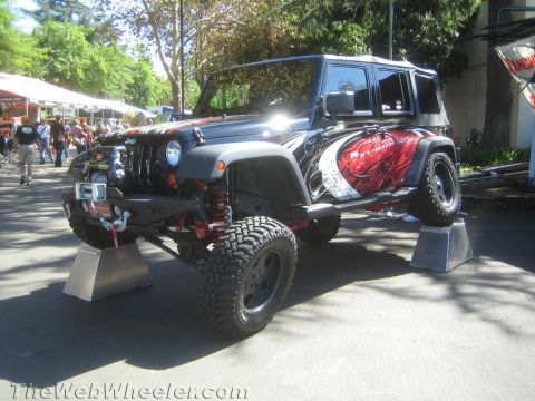 1990 Ford Bronco - black four-door Jeep Wrangler with red airbrushed graphics, a winch bumper and mud tires is raised on stands at an outdoor show.