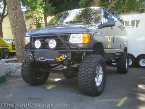 1990 Ford Bronco - lifted gray Ford van with a tube front bumper, off-road lights and mud tires sits beside a Kilby Enterprises trailer at a show.