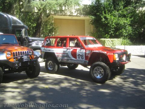 1990 Ford Bronco - red Jeep Cherokee race vehicle numbered 1718 with Jeepspeed and Goodyear decals sits parked beside an orange Jeep at a show.