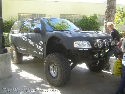 1990 Ford Bronco - matte black lifted SUV lettered American Trophy Truck sits on huge mud tires with off-road lights, parked outside near onlookers.