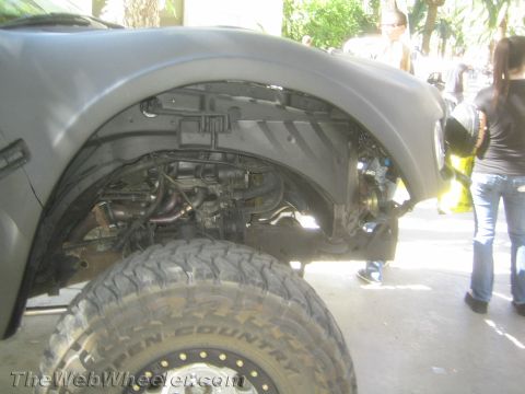 1990 Ford Bronco - close view inside the front wheel well of a matte gray lifted truck reveals long-travel suspension, axle and a mud tire.