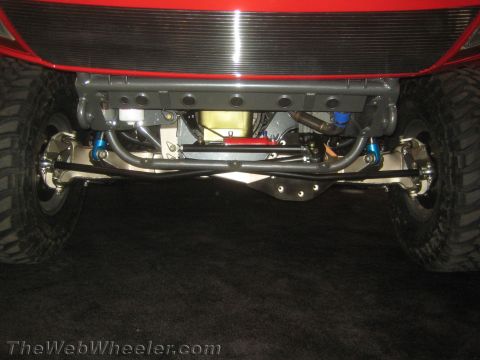 1990 Ford Bronco - underside of a red vehicle shows a polished front axle, blue-bodied shocks and tie rods over a dark carpeted display floor.