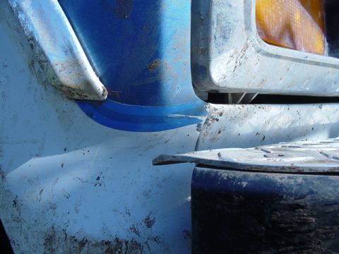 1985 Ford Bronco - Close-up view of the front corner of a damaged vehicle, revealing blue and white paint with visible dents and wear.