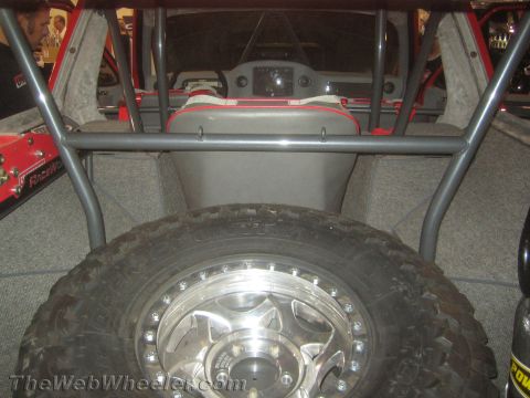 1990 Ford Bronco - Rear interior view of a caged off-road vehicle looking past gray seats to a mud tire mounted on a polished beadlock wheel.