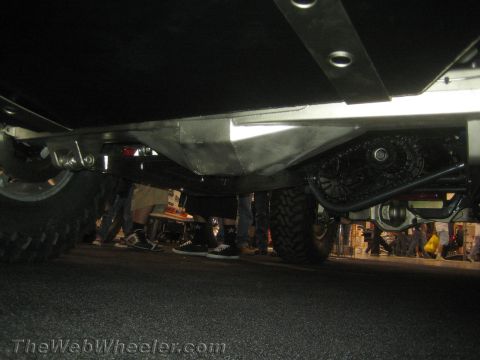 1990 Ford Bronco - Low underbody shot of an off-road vehicle on display showing the skid plate, axle, mud tires, and spectators' feet behind it.
