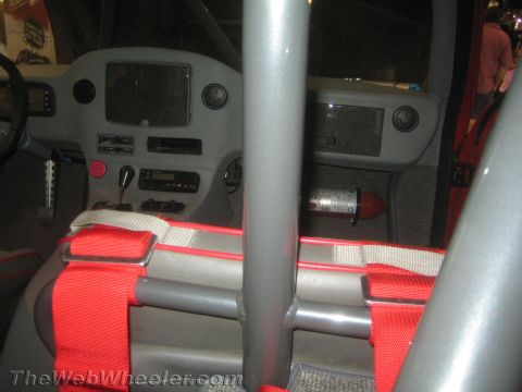 1990 Ford Bronco - Interior view of a caged off-road vehicle showing the gray dashboard, roll bar, red harness straps, and a small red fire extinguisher.