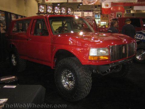 1990 Ford Bronco - Red lifted Ford full-size SUV with a billet grille and mud tires displayed at an indoor automotive show with overhead light bar.