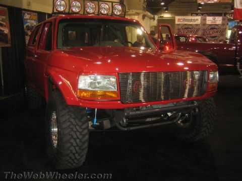 1990 Ford Bronco - Front three-quarter view of a red lifted Ford full-size SUV with billet grilles, round headlights, and a tube bumper at an indoor show.