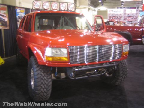1990 Ford Bronco - Front view of a red lifted Ford full-size SUV with a wide chrome billet grille and tube bumper on display at an indoor expo.
