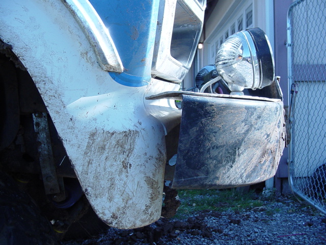 Front fender and bumper of a truck, covered in mud. A chrome headlight is visible