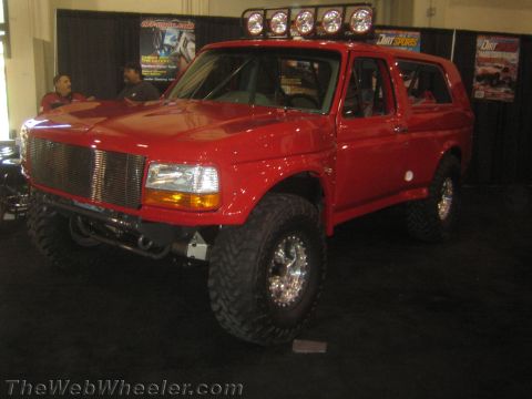1990 Ford Bronco - Front three-quarter view of a red lifted Ford full-size SUV with a roof-mounted light bar and billet grille at an indoor show.