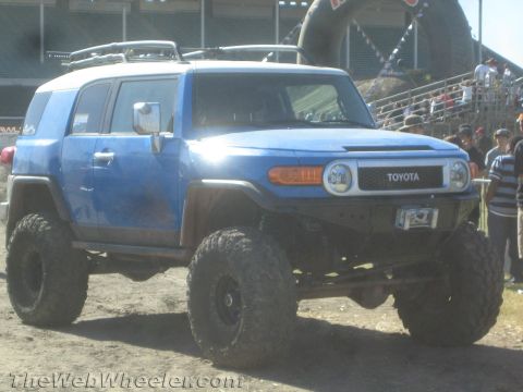 1990 Ford Bronco - Blue Toyota FJ Cruiser with white roof, lifted on mud tires and a tube bumper, at an outdoor off-road event with spectators.