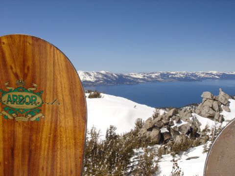 1990 Ford Bronco - A wooden snowboard with the Arbor logo is positioned in the foreground, overlooking a vast blue lake surrounded by snow-capped mountains.