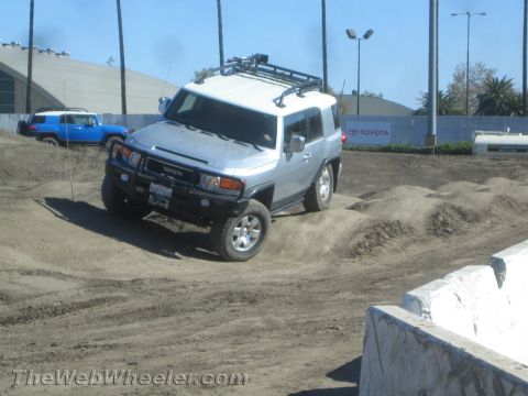 1990 Ford Bronco - Silver Toyota FJ Cruiser with roof rack climbing a graded dirt course in front of a Toyota banner, with a blue FJ in the background.