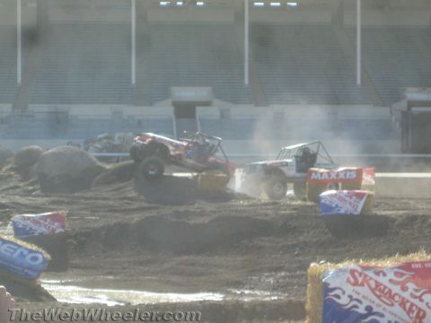 1990 Ford Bronco - Two tube-chassis off-road buggies churning dust as they climb obstacles on a dusty stadium competition course past sponsor hay bales.